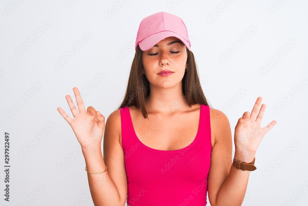 Young beautiful girl wearing pink casual t-shirt and cap over isolated white background relax and smiling with eyes closed doing meditation gesture with fingers. Yoga concept. Young beautiful girl wearing pink casual t-shirt and cap over isolated white background relax and smiling with eyes closed doing meditation gesture with fingers. Yoga concept.