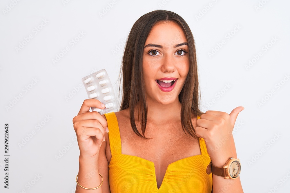 Young beautiful girl holding medicine pills standing over isolated white background pointing and showing with thumb up to the side with happy face smiling Young beautiful girl holding medicine pills standing over isolated white background pointing and showing with thumb up to the side with happy face smiling