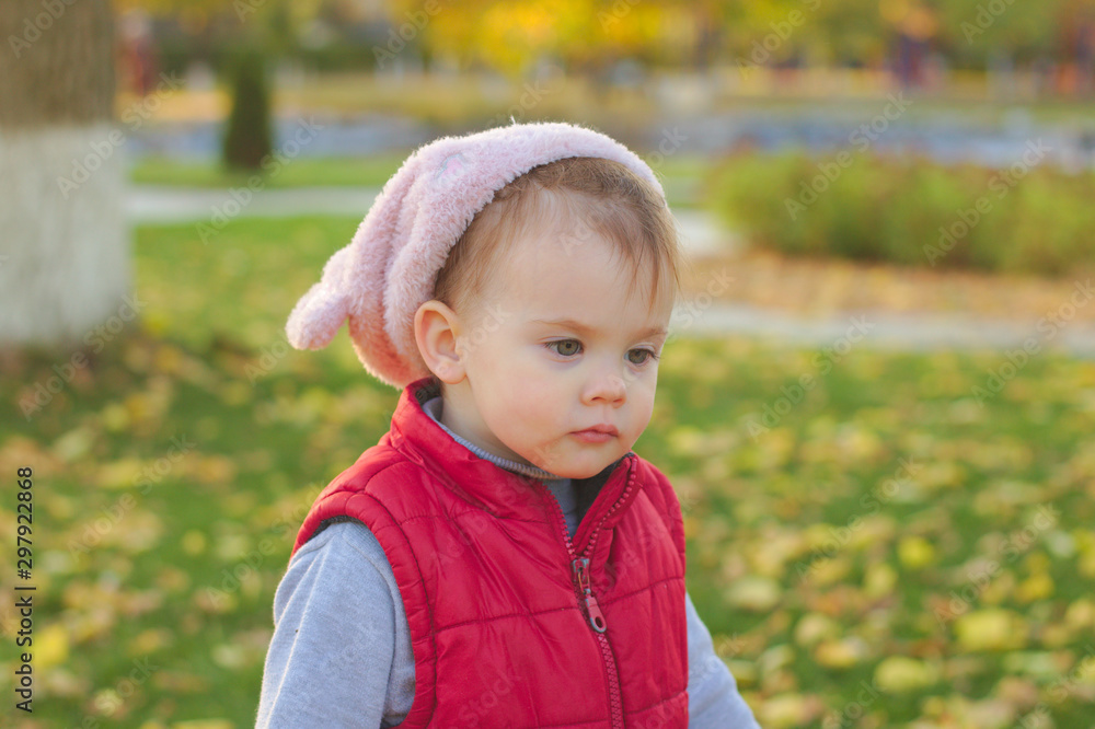 A small child in a fluffy pink hat and red vest is running in the autumn park. Beautiful fall sunny day outdoors.