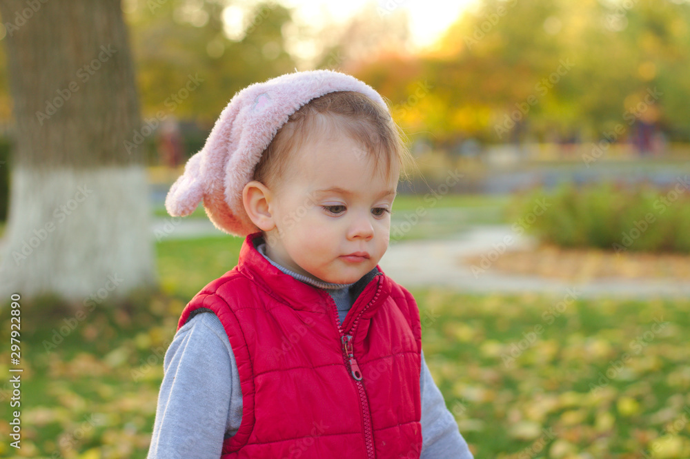 A small child in a fluffy pink hat and red vest is running in the autumn park. Beautiful fall sunny day outdoors. A small child in a fluffy pink hat and red vest is running in the autumn park. Beautiful fall sunny day outdoors.