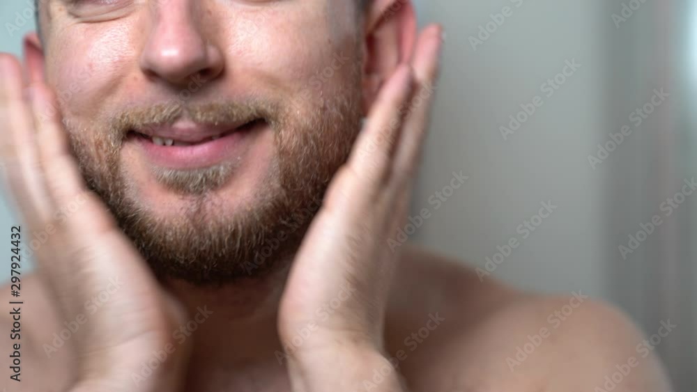 Young handsome man smoothing his cutted and cleaned bear with hands in front of a mirror. Man doing morning skincare procedures in the bathroom. Successful businessman brushing his beard before going Young handsome man smoothing his cutted and cleaned bear with hands in front of a mirror. Man doing morning skincare procedures in the bathroom. Successful businessman brushing his beard before going