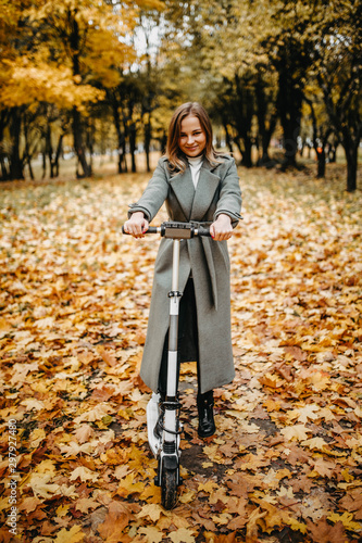Woman in a coat in autumn on an electric scooter in an autumn park. Riding on electric vehicles in cold weather.