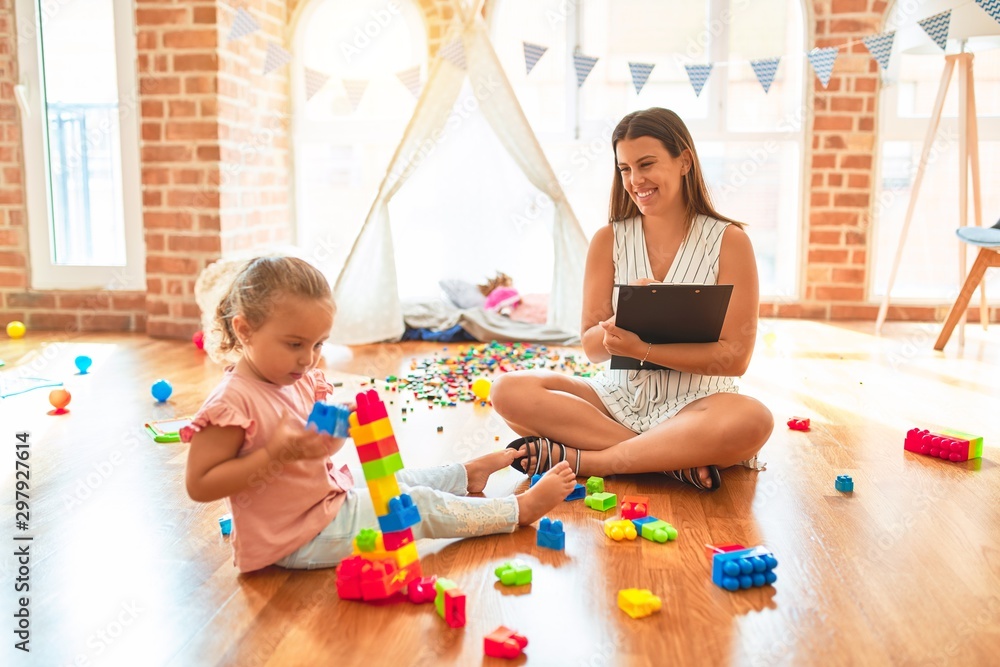 Fototapeta premium Beautiful psycologist and blond toddler girl doing therapy building tower using plastic blocks at kindergarten