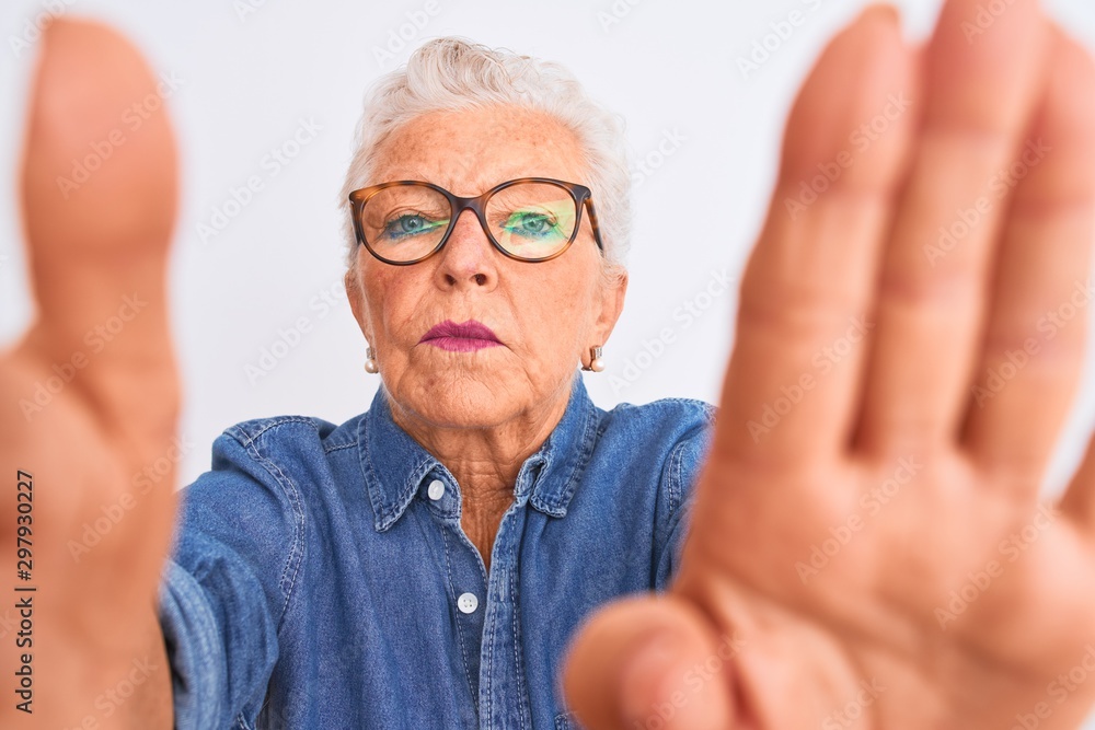 Grey-haired woman wearing denim shirt and glasses make selfie over isolated white background with open hand doing stop sign with serious and confident expression, defense gesture Grey-haired woman wearing denim shirt and glasses make selfie over isolated white background with open hand doing stop sign with serious and confident expression, defense gesture