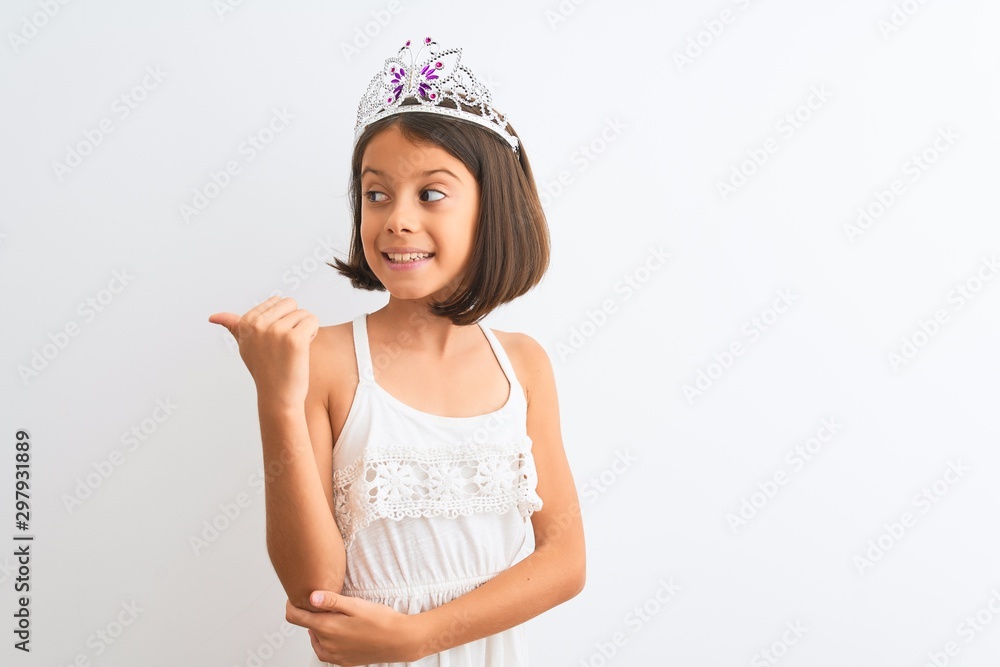 Beautiful child girl wearing princess crown standing over isolated white background smiling with happy face looking and pointing to the side with thumb up. Beautiful child girl wearing princess crown standing over isolated white background smiling with happy face looking and pointing to the side with thumb up.