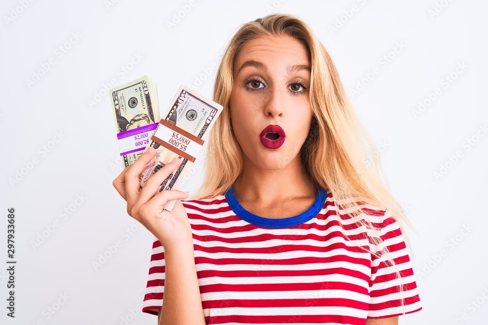 Beautiful woman wearing red striped t-shirt holding dollars over isolated white background scared in shock with a surprise face, afraid and excited with fear expression Beautiful woman wearing red striped t-shirt holding dollars over isolated white background scared in shock with a surprise face, afraid and excited with fear expression