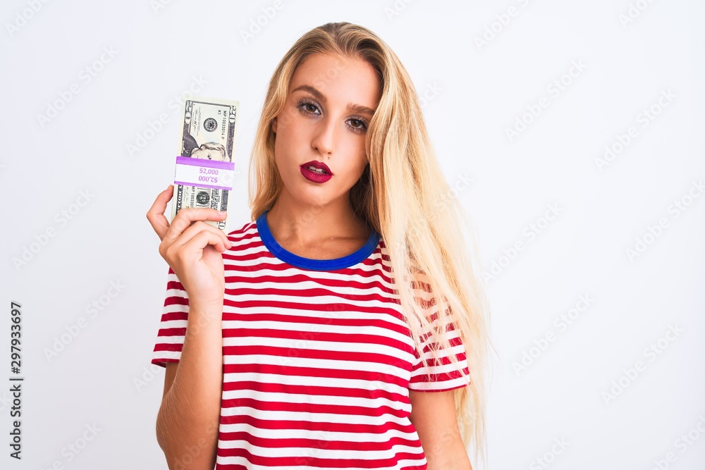 Beautiful woman wearing red striped t-shirt holding dollars over isolated white background with a confident expression on smart face thinking serious Beautiful woman wearing red striped t-shirt holding dollars over isolated white background with a confident expression on smart face thinking serious