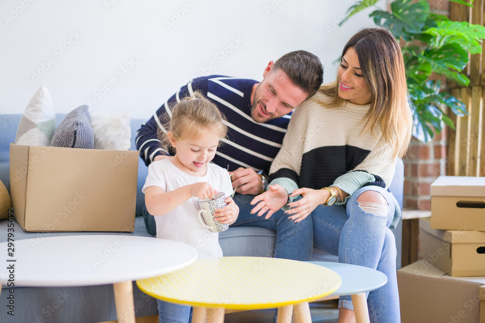 Beautiful family, parents sitting on the sofa drinking coffee looking his kid playing at new home around cardboard boxes Beautiful family, parents sitting on the sofa drinking coffee looking his kid playing at new home around cardboard boxes
