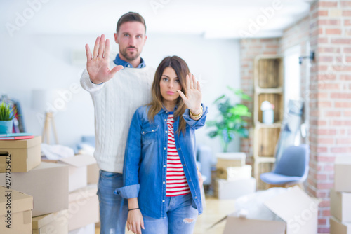 Young beautiful couple standing at new home around cardboard boxes doing stop sing with palm of the hand. Warning expression with negative and serious gesture on the face.