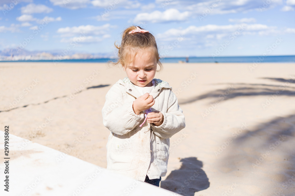Beautiful toddler child girl wearing jacket playing with the sand on the beach Beautiful toddler child girl wearing jacket playing with the sand on the beach