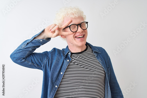 Young albino blond man wearing denim shirt and glasses over isolated white background Doing peace symbol with fingers over face, smiling cheerful showing victory