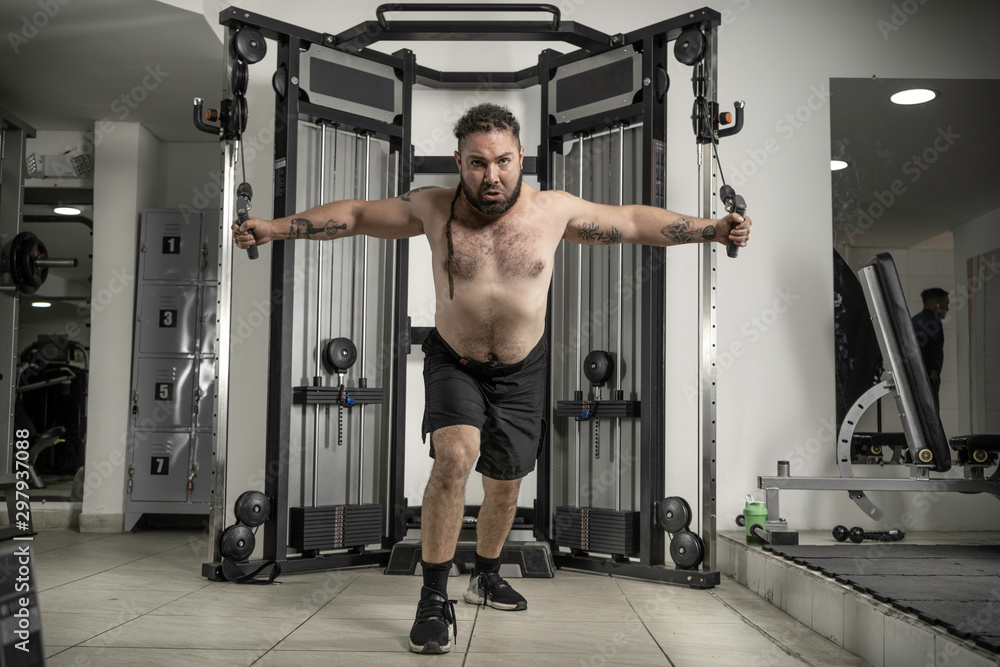 Fototapeta premium Young man in T-shirt at gym with rasta hair working triceps, chest, back, arms with pulley
