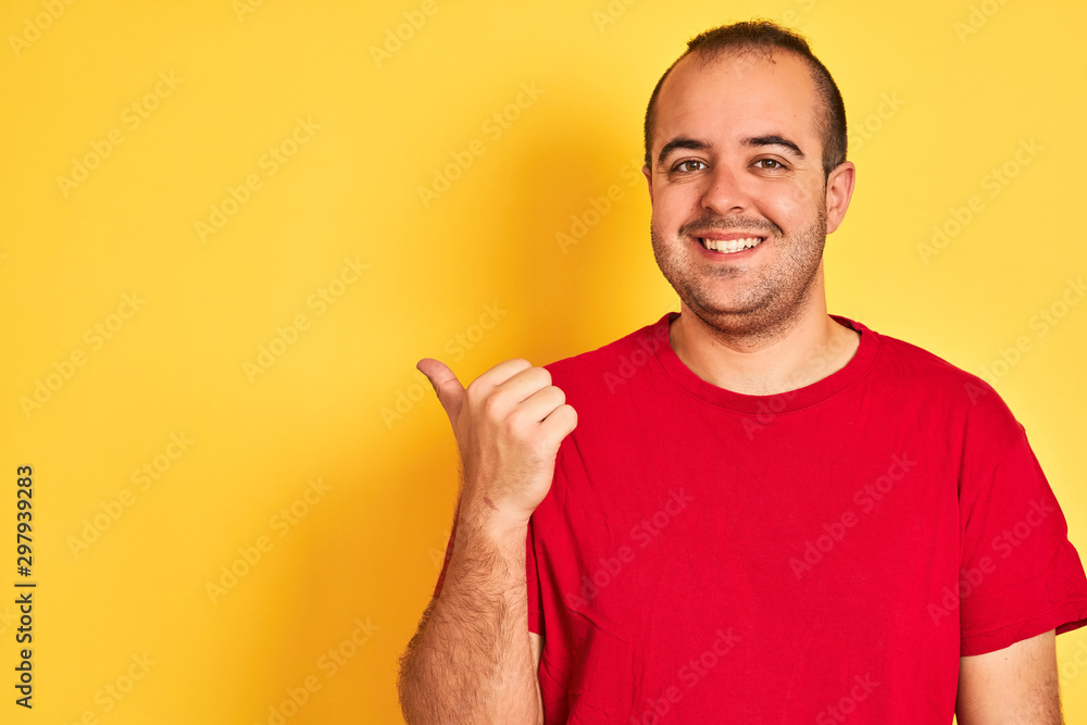 Young man wearing red casual t-shirt standing over isolated yellow background pointing and showing with thumb up to the side with happy face smiling Young man wearing red casual t-shirt standing over isolated yellow background pointing and showing with thumb up to the side with happy face smiling
