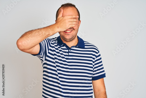 Young man wearing casual striped polo standing over isolated white background smiling and laughing with hand on face covering eyes for surprise. Blind concept.