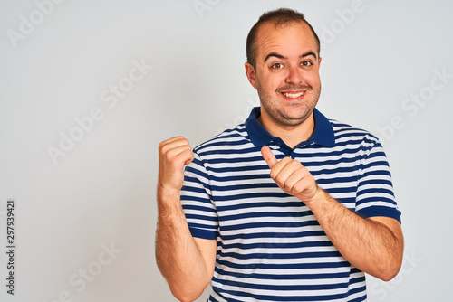 Young man wearing casual striped polo standing over isolated white background Pointing to the back behind with hand and thumbs up, smiling confident