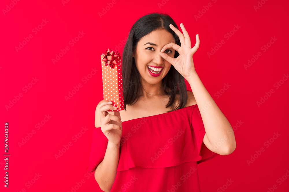 Young beautiful chinese woman holding valentine gift standing over isolated red background with happy face smiling doing ok sign with hand on eye looking through fingers Young beautiful chinese woman holding valentine gift standing over isolated red background with happy face smiling doing ok sign with hand on eye looking through fingers