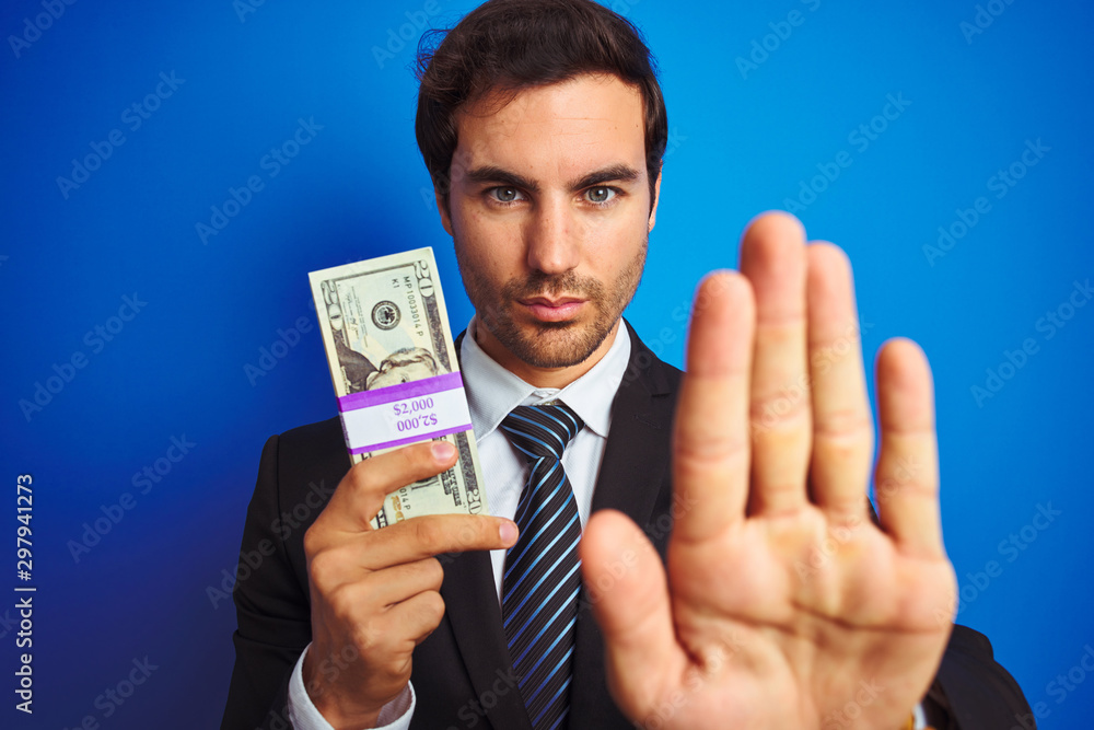 Young handsome businessman wearing suit holding dollars over isolated blue background with open hand doing stop sign with serious and confident expression, defense gesture Young handsome businessman wearing suit holding dollars over isolated blue background with open hand doing stop sign with serious and confident expression, defense gesture