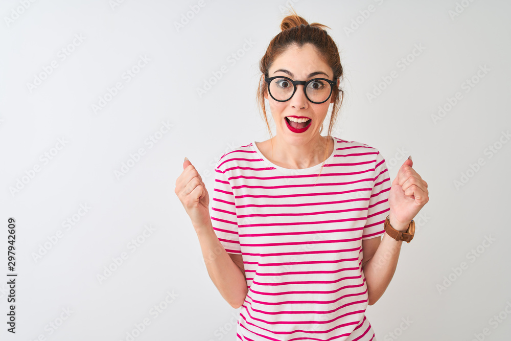 Redhead woman wearing glasses striped t-shirt and pigtail over isolated white background celebrating surprised and amazed for success with arms raised and open eyes. Winner concept. Redhead woman wearing glasses striped t-shirt and pigtail over isolated white background celebrating surprised and amazed for success with arms raised and open eyes. Winner concept.