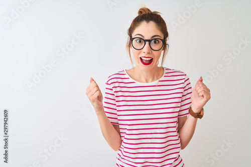 Redhead woman wearing glasses striped t-shirt and pigtail over isolated white background celebrating surprised and amazed for success with arms raised and open eyes. Winner concept.