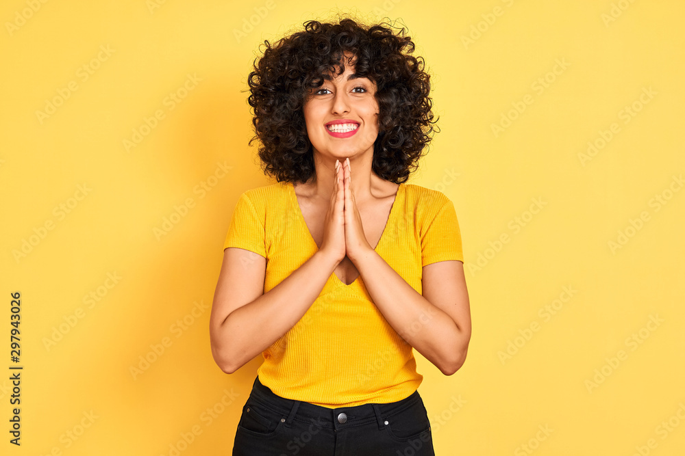 Young arab woman with curly hair wearing t-shirt standing over isolated yellow background praying with hands together asking for forgiveness smiling confident. Young arab woman with curly hair wearing t-shirt standing over isolated yellow background praying with hands together asking for forgiveness smiling confident.
