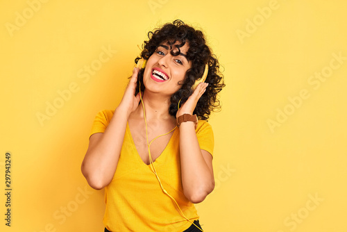 Arab woman with curly hair listening to music using headphones over isolated yellow background with a happy and cool smile on face. Lucky person.