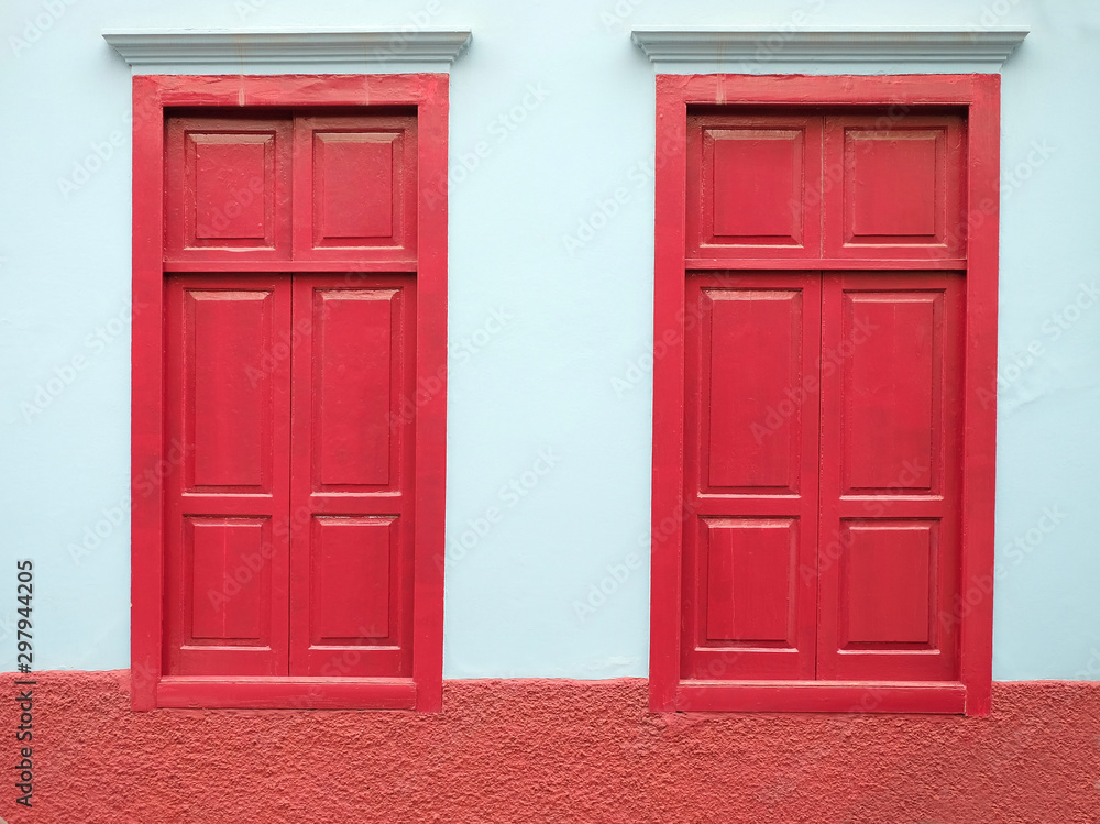 Two burgundy red closed windows and a pale blue wall, small town architecture detail. Two burgundy red closed windows and a pale blue wall, small town architecture detail.