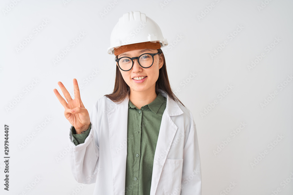 Young chinese engineer woman wearing coat helmet glasses over isolated white background showing and pointing up with fingers number three while smiling confident and happy. Young chinese engineer woman wearing coat helmet glasses over isolated white background showing and pointing up with fingers number three while smiling confident and happy.