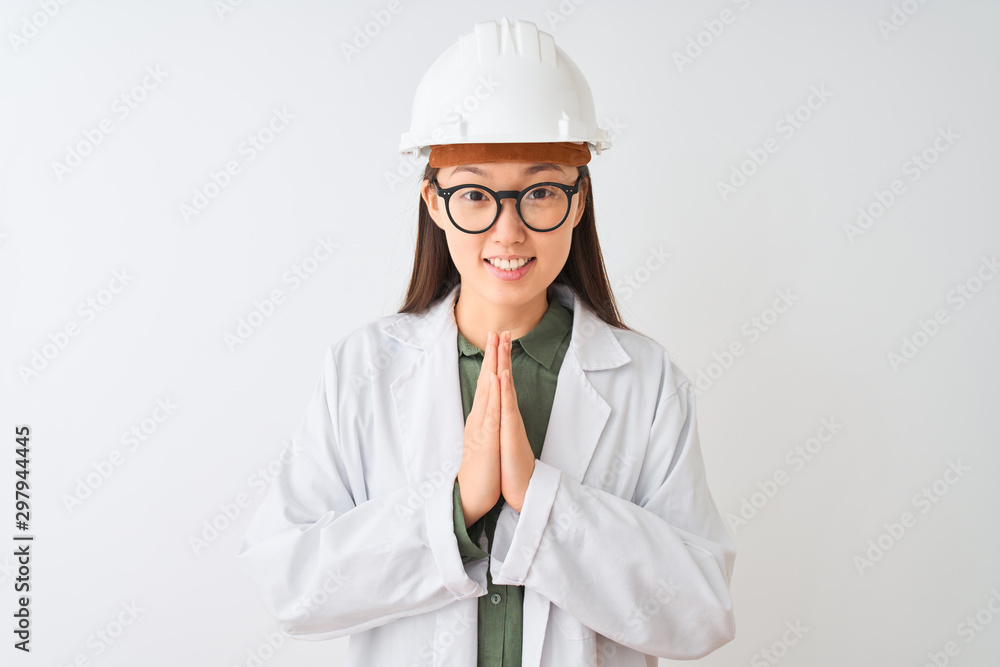 Young chinese engineer woman wearing coat helmet glasses over isolated white background praying with hands together asking for forgiveness smiling confident. Young chinese engineer woman wearing coat helmet glasses over isolated white background praying with hands together asking for forgiveness smiling confident.