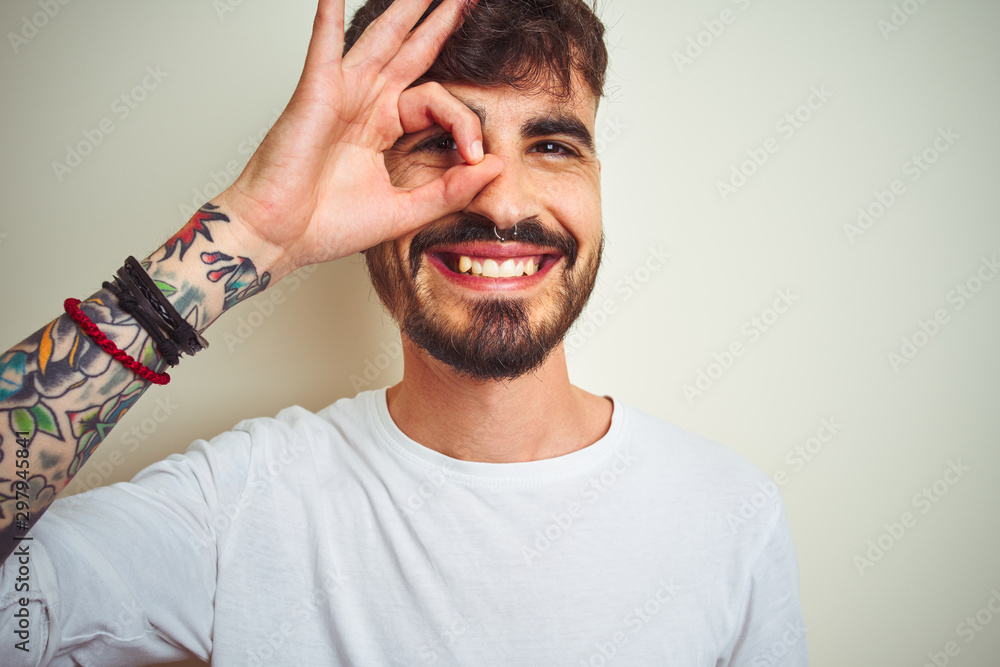 Young man with tattoo wearing t-shirt standing over isolated white background doing ok gesture with hand smiling, eye looking through fingers with happy face. Young man with tattoo wearing t-shirt standing over isolated white background doing ok gesture with hand smiling, eye looking through fingers with happy face.