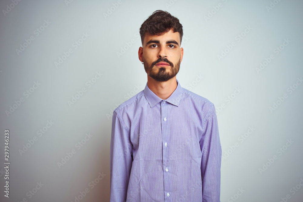 Obraz premium Young man with tattoo wearing purple shirt standing over isolated white background with serious expression on face. Simple and natural looking at the camera.