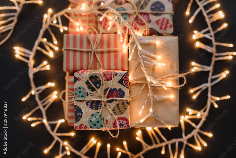 Fototapeta premium A bunch of Christmas gift boxes in multicolored kraft wrap under the garland with warm light. Selective focus on the top gift. Preparation for celebrating. Merry Christmas and Happy New Year concept.