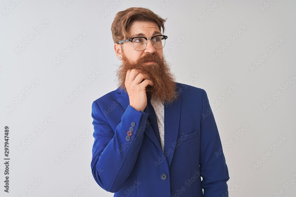 Young redhead irish businessman wearing suit and glasses over isolated white background with hand on chin thinking about question, pensive expression. Smiling with thoughtful face. Doubt concept.