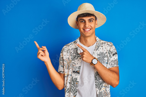 Indian man on vacation wearing hawiaian shirt summer hat over isolated blue background smiling and looking at the camera pointing with two hands and fingers to the side.