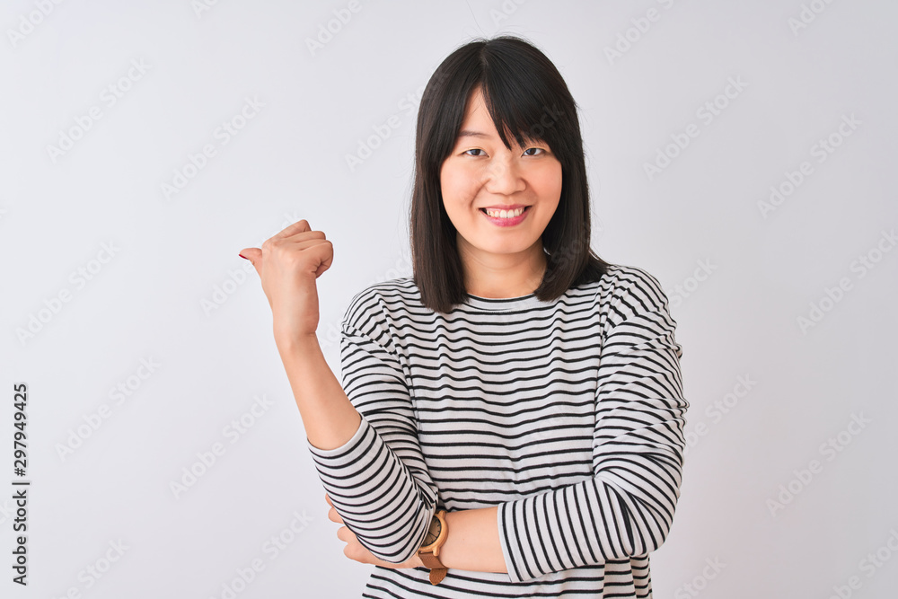 Young beautiful chinese woman wearing black striped t-shirt over isolated white background smiling with happy face looking and pointing to the side with thumb up. Young beautiful chinese woman wearing black striped t-shirt over isolated white background smiling with happy face looking and pointing to the side with thumb up.