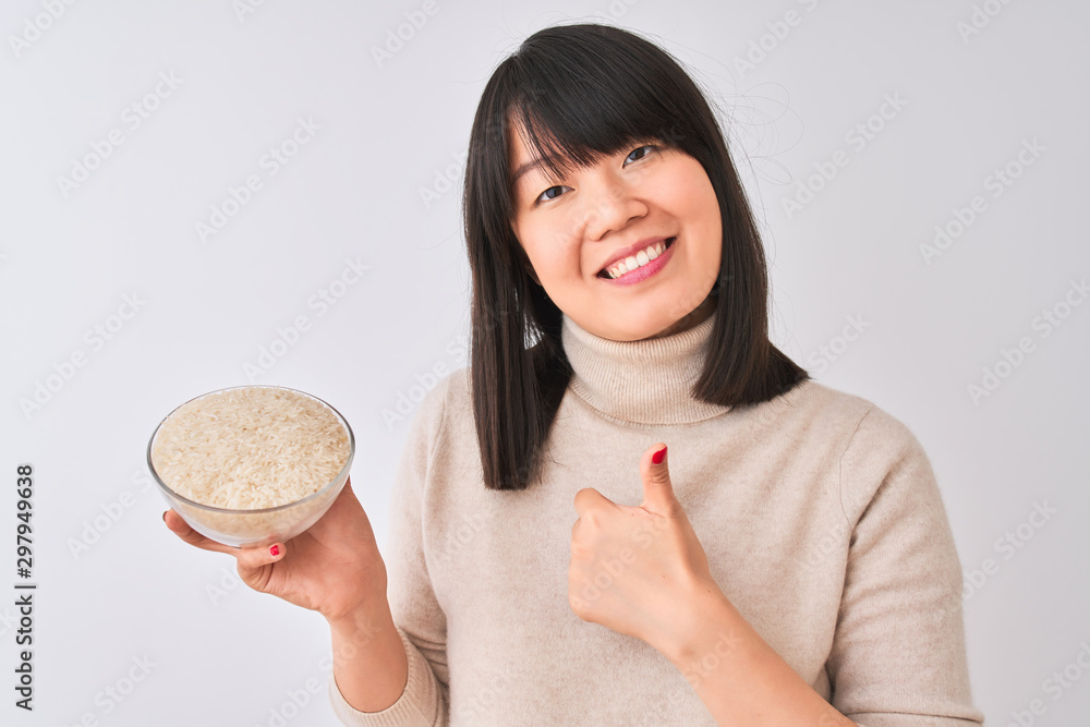 Young beautiful Chinese woman holding bowl with rice over isolated white background happy with big smile doing ok sign, thumb up with fingers, excellent sign Young beautiful Chinese woman holding bowl with rice over isolated white background happy with big smile doing ok sign, thumb up with fingers, excellent sign