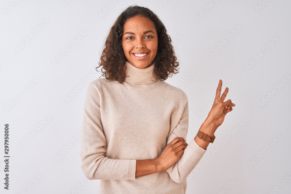 Young brazilian woman wearing turtleneck sweater standing over isolated white background smiling with happy face winking at the camera doing victory sign. Number two.