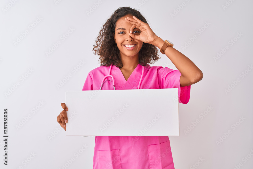 Young brazilian nurse woman holding banner standing over isolated white background with happy face smiling doing ok sign with hand on eye looking through fingers Young brazilian nurse woman holding banner standing over isolated white background with happy face smiling doing ok sign with hand on eye looking through fingers
