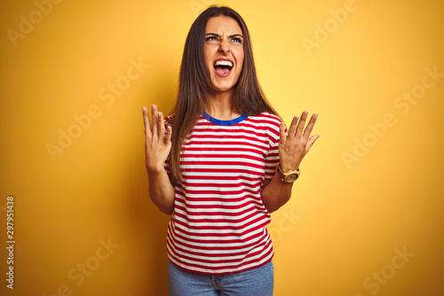 Young beautiful woman wearing striped t-shirt standing over isolated yellow background crazy and mad shouting and yelling with aggressive expression and arms raised. Frustration concept.