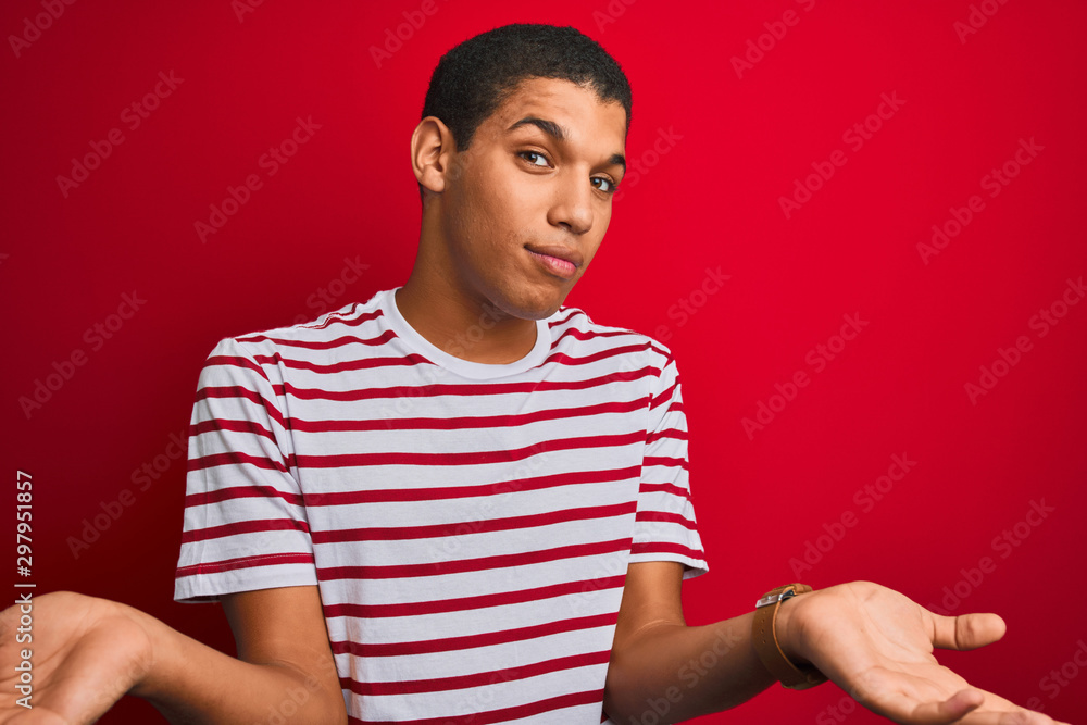 Young handsome arab man wearing striped t-shirt over isolated red background clueless and confused with open arms, no idea concept. Young handsome arab man wearing striped t-shirt over isolated red background clueless and confused with open arms, no idea concept.