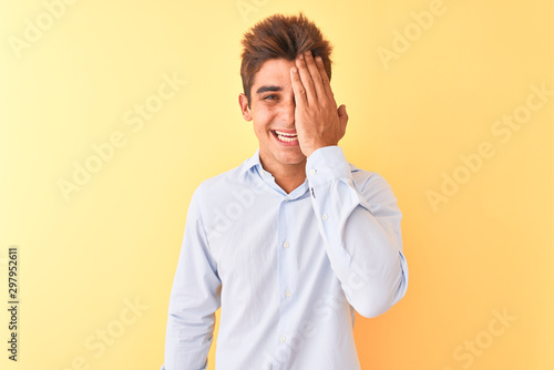 Young handsome businessman wearing elegant shirt over isolated yellow background covering one eye with hand, confident smile on face and surprise emotion.