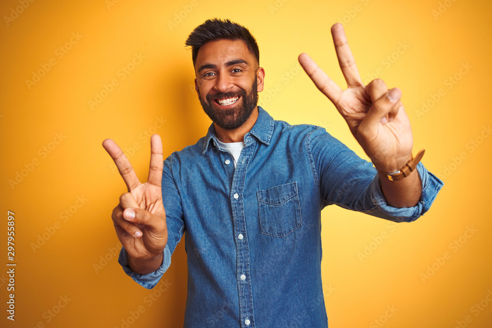 Young indian man wearing denim shirt standing over isolated yellow background smiling looking to the camera showing fingers doing victory sign. Number two. Young indian man wearing denim shirt standing over isolated yellow background smiling looking to the camera showing fingers doing victory sign. Number two.