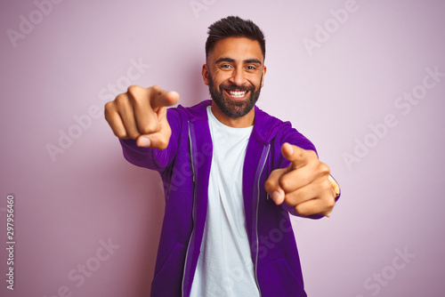 Young indian man wearing purple sweatshirt standing over isolated pink background pointing to you and the camera with fingers, smiling positive and cheerful