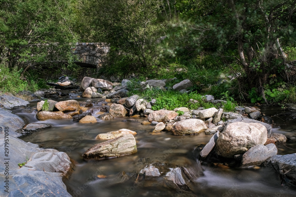 Ancient bridge over a Mountain Creek. Mountain river in the forest on green trees background. Forest landscape with water movement between large stones. Long exposure. Ancient bridge over a Mountain Creek. Mountain river in the forest on green trees background. Forest landscape with water movement between large stones. Long exposure.