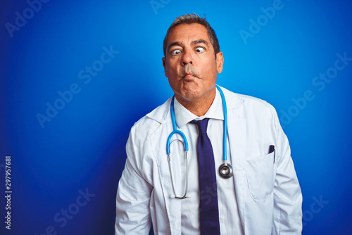 Handsome middle age doctor man wearing stethoscope over isolated blue background making fish face with lips, crazy and comical gesture. Funny expression.