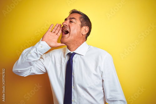 Handsome middle age businessman standing over isolated yellow background shouting and screaming loud to side with hand on mouth. Communication concept.