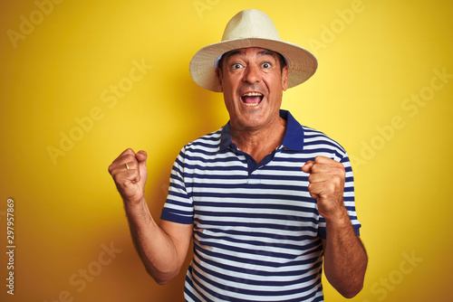 Handsome middle age man wearing striped polo and hat over isolated yellow background very happy and excited doing winner gesture with arms raised, smiling and screaming for success. 