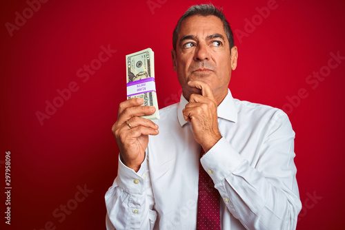 Handsome middle age businessman holding dollars over isolated red background serious face thinking about question, very confused idea
