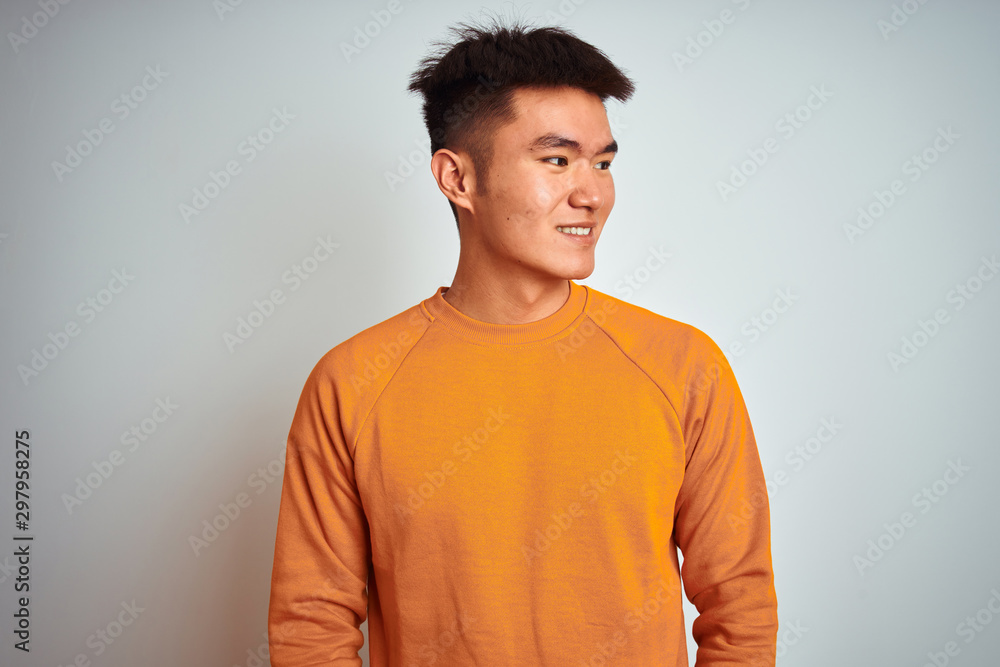 Young asian chinese man wearing orange sweater standing over isolated white background looking away to side with smile on face, natural expression. Laughing confident. Young asian chinese man wearing orange sweater standing over isolated white background looking away to side with smile on face, natural expression. Laughing confident.