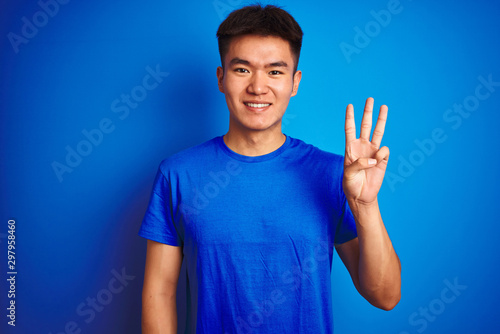 Young asian chinese man wearing t-shirt standing over isolated blue background showing and pointing up with fingers number three while smiling confident and happy.