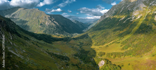 AERIAL; panorama of steep stony slopes of highland; melting snow in summer sunny evening; beautiful natural landscape with mountain ridges and river valley; high dangerous rocky tops of Caucasus